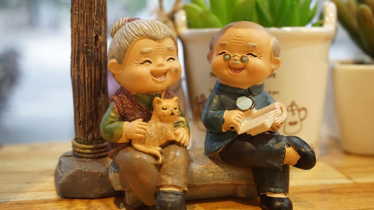 Zdjęcie przedstawia figurkę uśmiechniętej babci trzymającej na kolanach kota oraz siedzącego obok niej uśmiechniętego dziadka trzymającego lupę i książkę. 