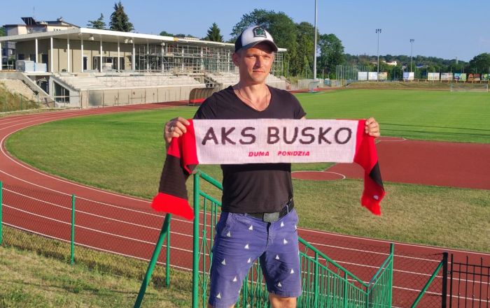 Na zdjęciu mężczyzna trzyma w rękach szalik kibica AKS Busko. W tle bieżnia, murawa i zabudowania buskiego stadionu
