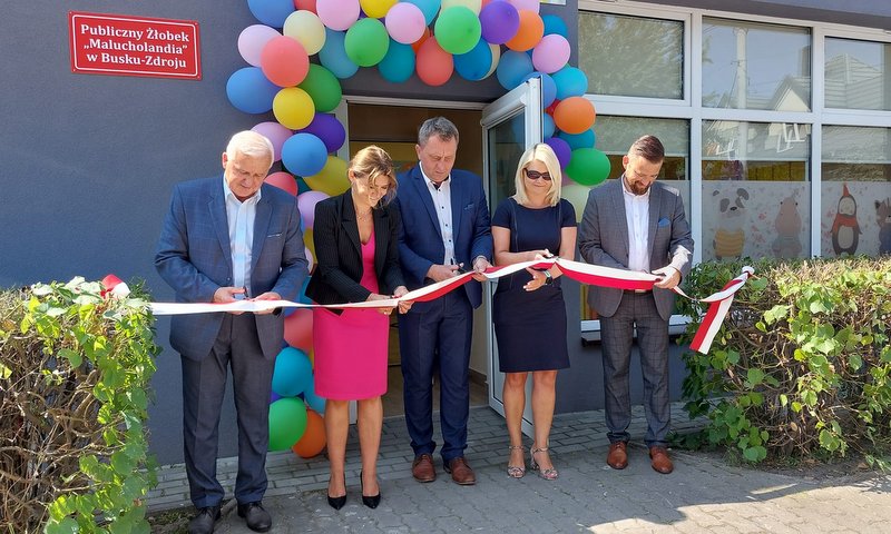 Pięcioro elegancko ubranych osób przecina wstęgę. W tle udekorowany kolorowymi balonami budynek