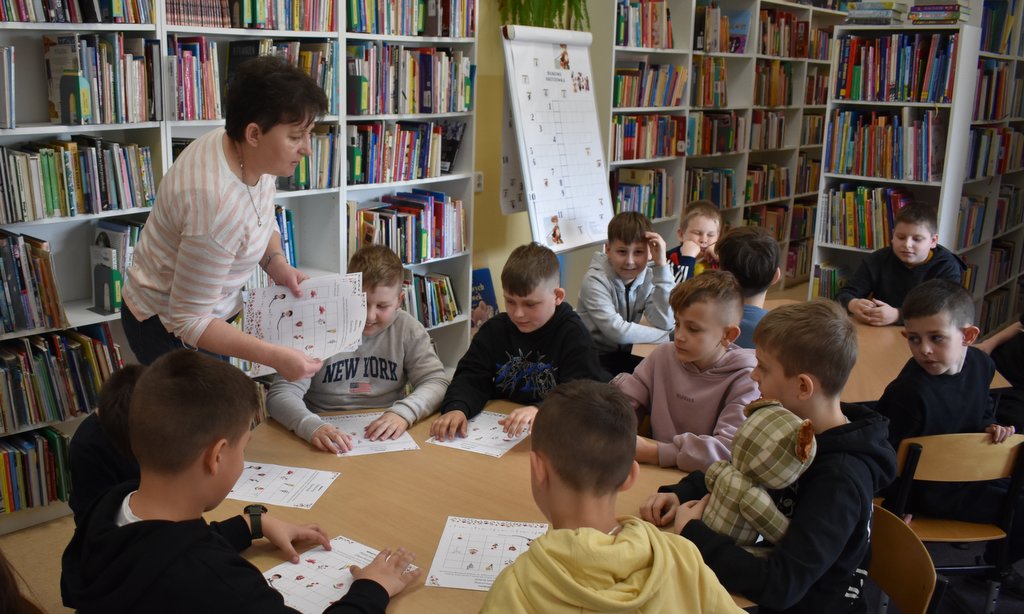Kobieta rozdaje karty pracy dzieciom, w tle regały biblioteczne