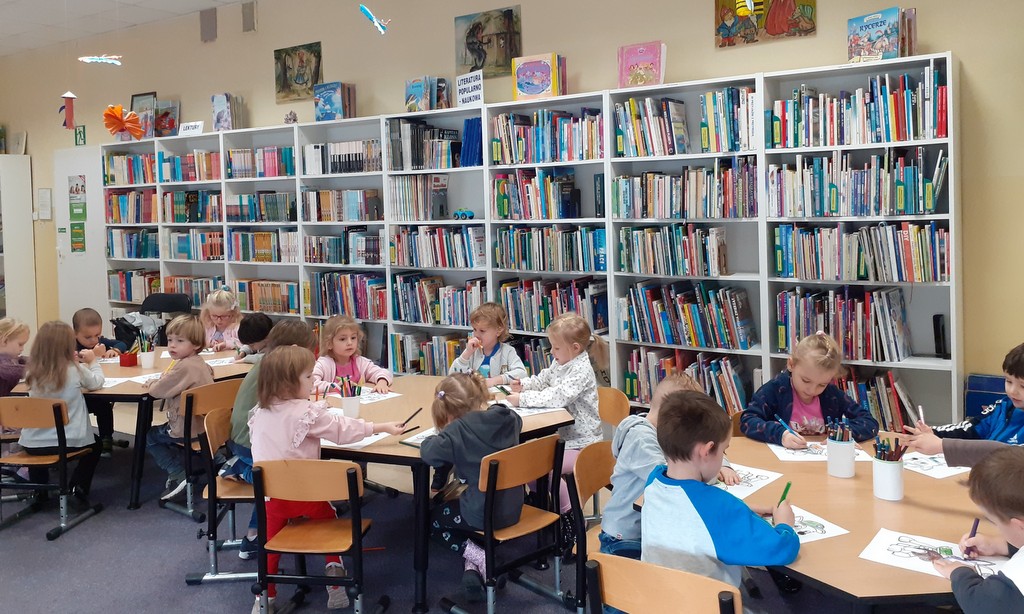 Dzieci siedzą przy trzech stoliczkach i rysują w tle widać regały z książkami