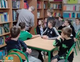 Mężczyzna prowadzący spotkanie przegląda regał z książkami  w otoczeniu dzieci Mężczyzna prowadzący spotkanie przegląda regał z książkami  w otoczeniu dzieci