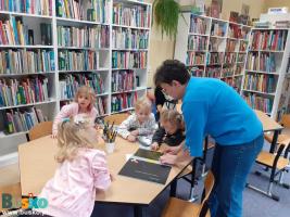 Dzieci przy stoliku, obok stoi kobieta, w tle regał z książkami