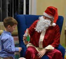 Mikoaj-siedzi-w-fotelu-i-daje-maemu-chopcu-w-niebieskiej-koszuli-prezent