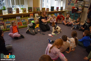 Kobieta ma na dłoni pacynkę, a pies siedzi przed nią. Wokół siedzą dzieci