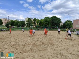 Zdjęcie przedstawia dziewięciu zawodników, czterech piłkarzy z jednej drużyny (pomarańczowe stroje)                          i czterech z drugiej (białe stroje). Na zdjęciu w oddali znajduje się bramk