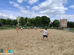 Zdjęcie przedstawia 10 zawodników. Czterech w strojach pomarańczowych i czterech w strojach biało-czarnych. W bramkach bramkarze drużyn, a w oddali boiska sędzia sędziujący mecz. Jeden z bramkarzy wyr