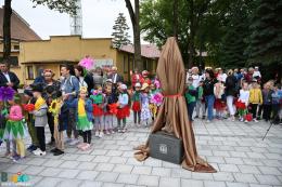 Rzeźba osłonięta materiałem gotowa do odsłonięcia, w tle przedszkolaki w kwiecistych strojach