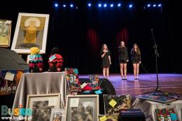 Trwa część artystyczna - na scenie trzy osoby, na pierwszym planie widac gadżety na llicyatcję